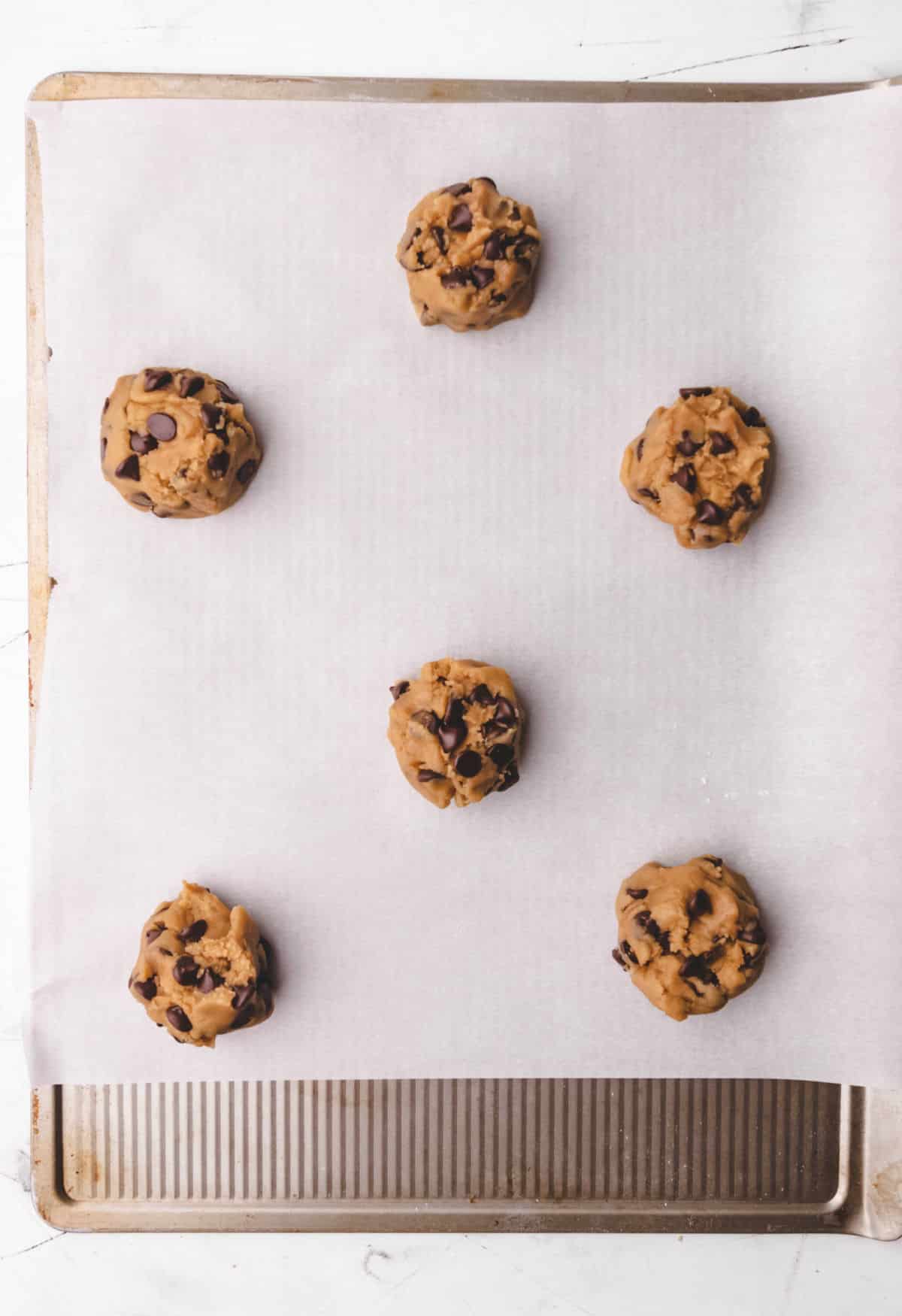 Six chocolate chip cookie dough balls on a baking sheet. 