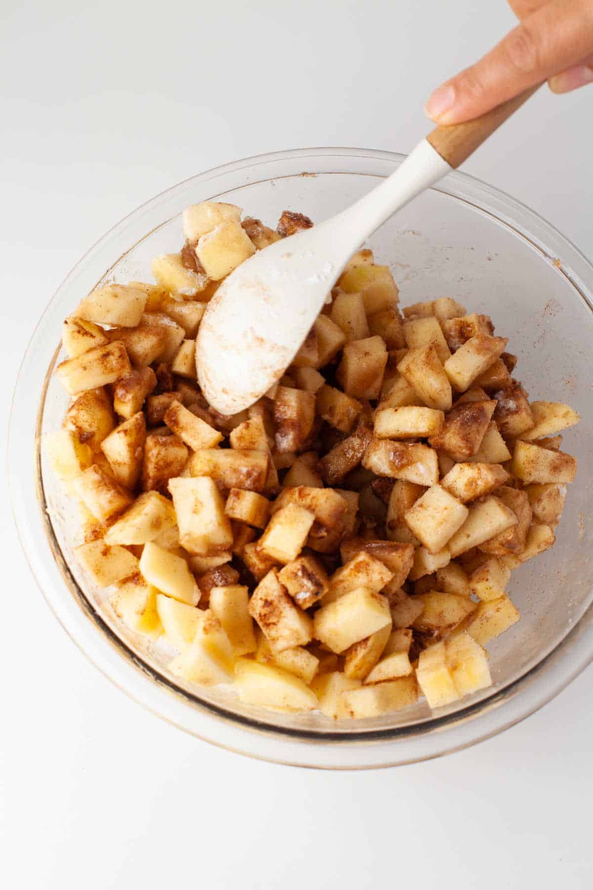 A wooden spoon stirring together apple pie filling in a glass mixing bowl. 