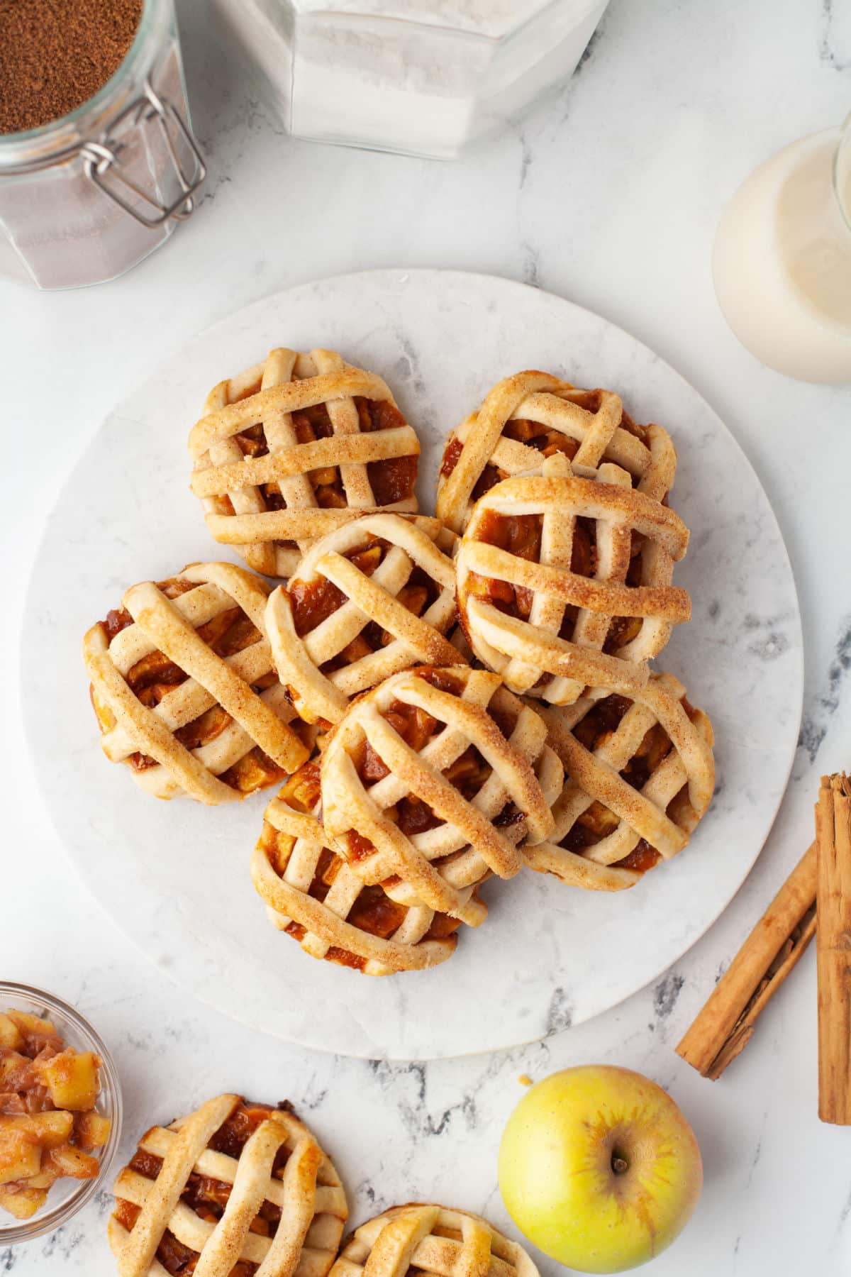 Stack of apple pie cookies surrounded by cinnamon sticks apples and flour cannister. 