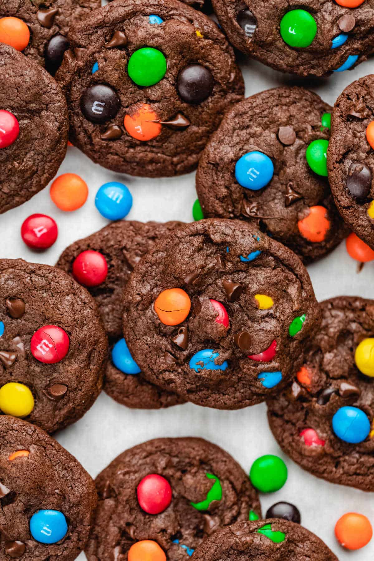 Overlapping rows of chocolate M&M cookies surrounded by M&Ms on parchment paper. 