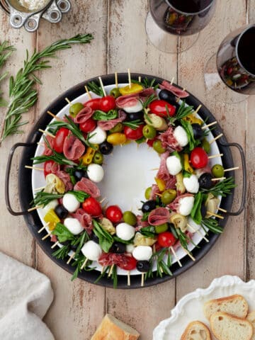 A charcuterie wreath surrounded by glasses of wine and sliced baguette.