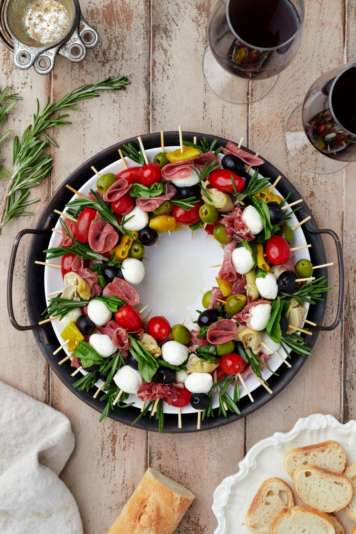 A charcuterie wreath surrounded by glasses of wine and sliced baguette.