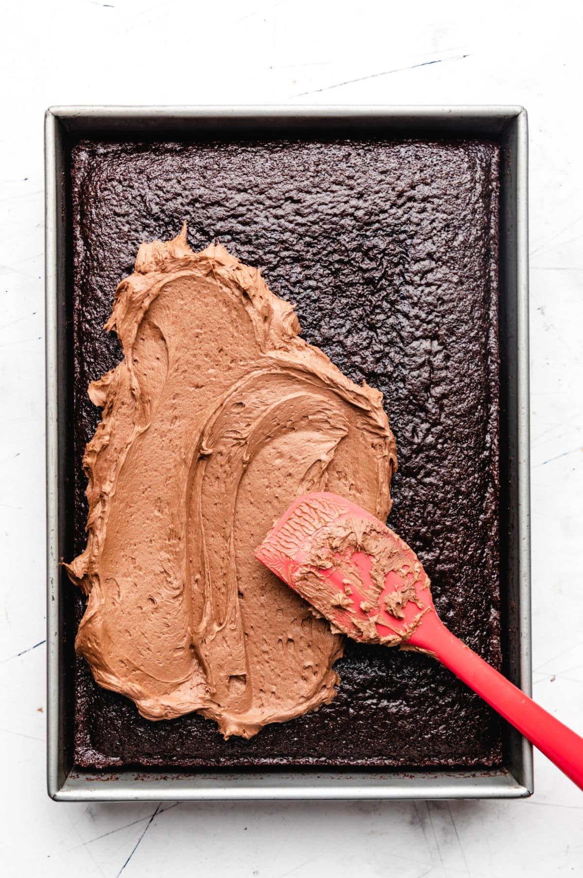 A red spatula spreading chocolate buttercream frosting on a chocolate sheet cake.