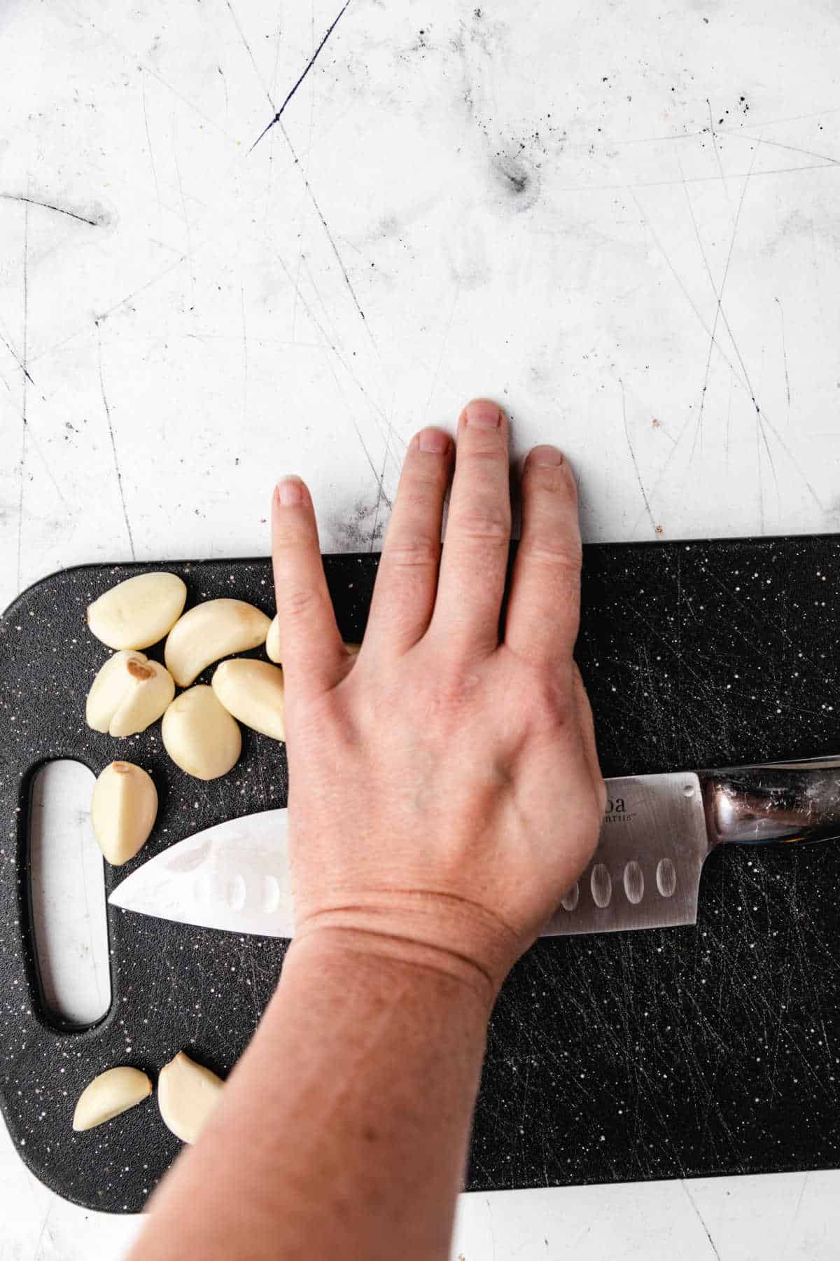 A hand pushing down on a knife to smash cloves of garlic. 