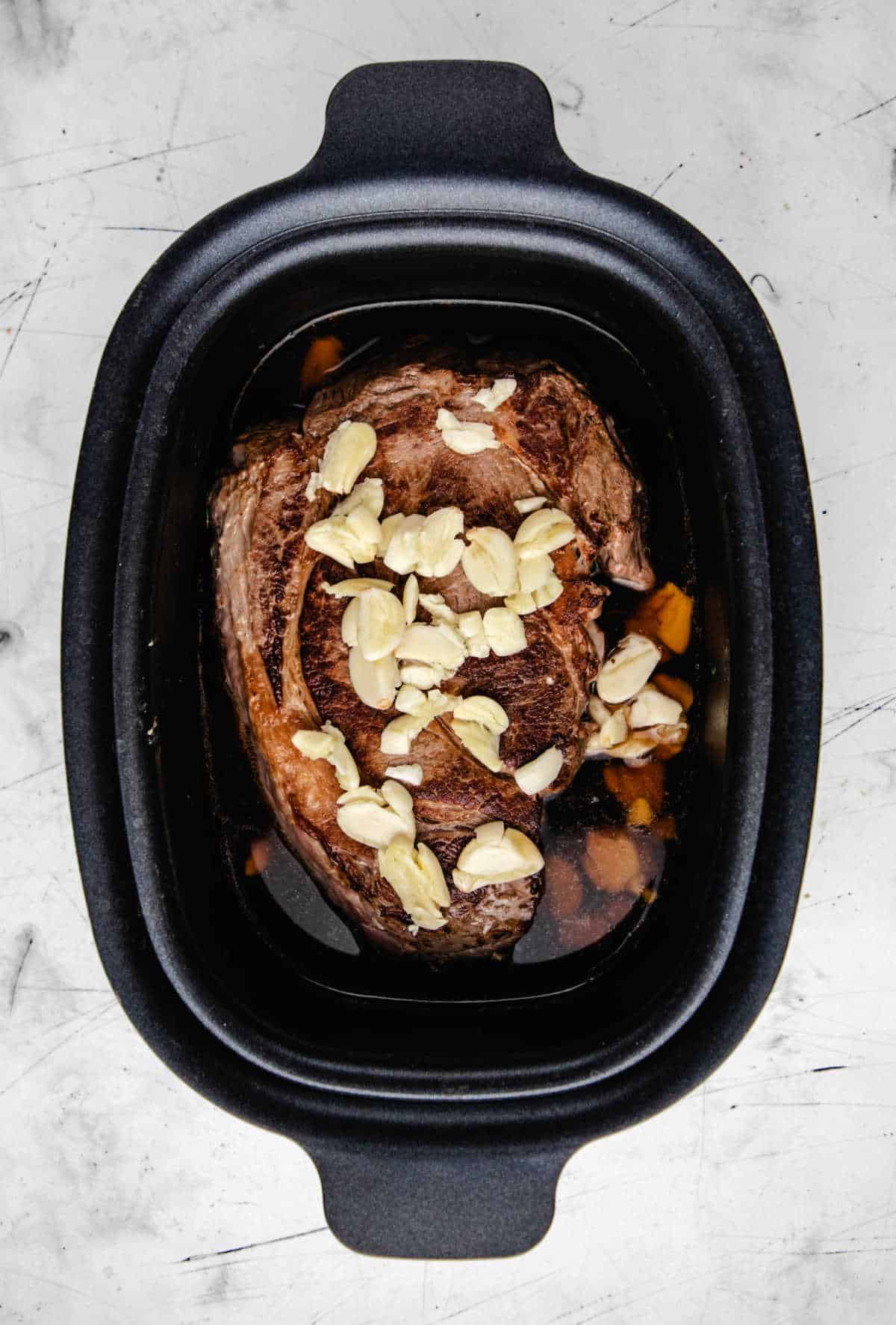 Browned chuck roast topped with cloves of garlic in a slow cooker.