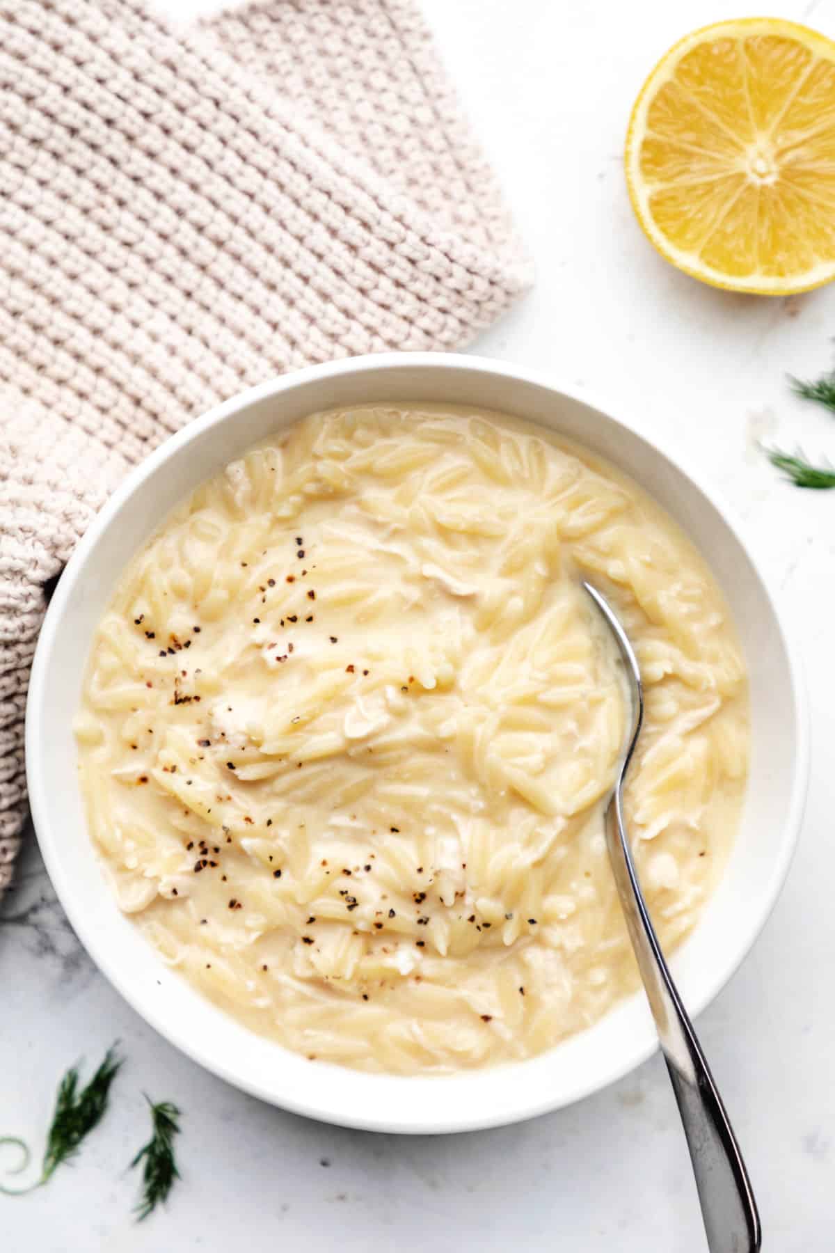 A bowl of avgolemono soup with a silver spoon in it next to a sliced lemon.