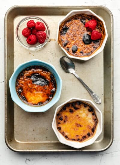 Three dishes of creme bruhlee on a baking sheet with a spoon and a dish of raspberries.