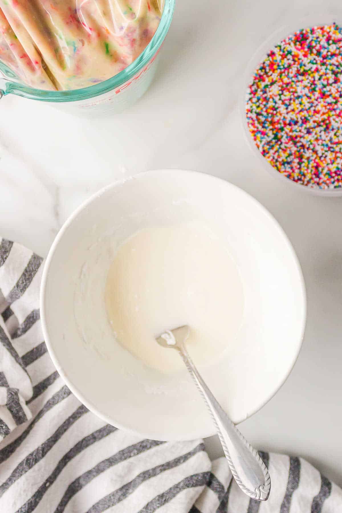 Donut glaze in a mixing bowl next to a dish of sprinkles. 