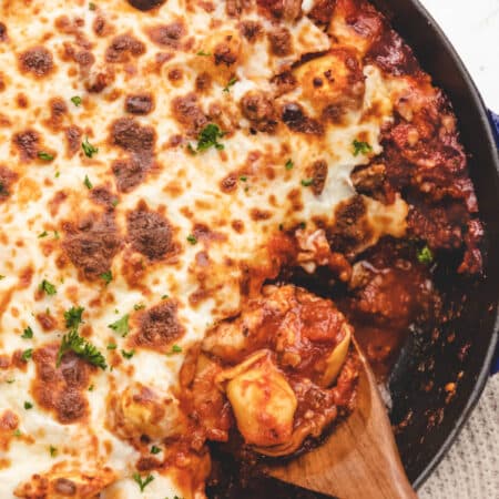 A wooden spoon scooping up baked tortellini from a skillet.