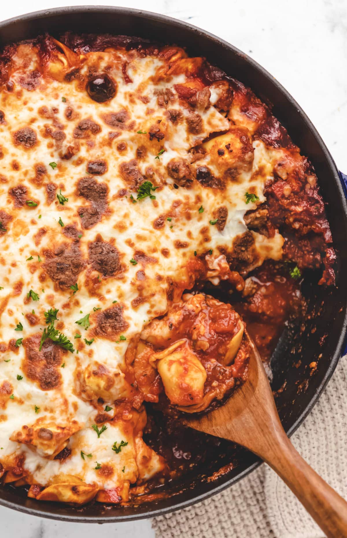 A wooden spoon scooping up baked tortellini from a skillet.