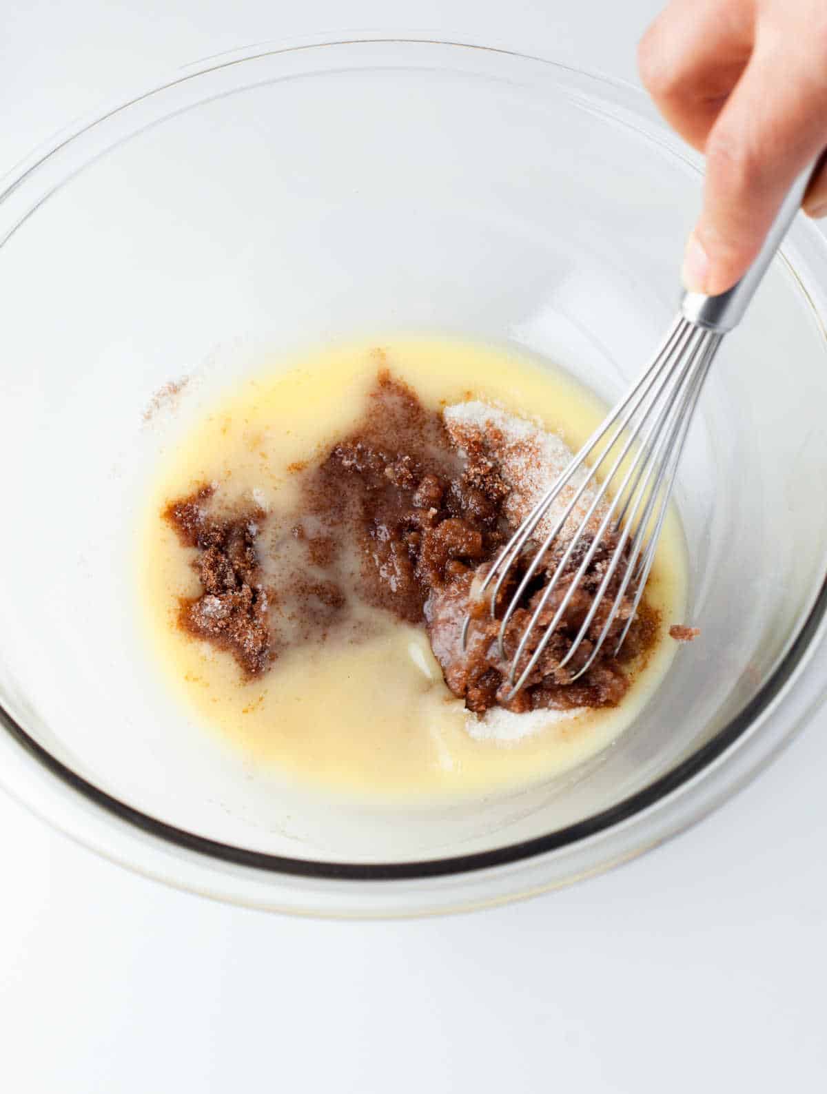 A whisk stirring melted butter and sugars together in a glass mixing bowl. 