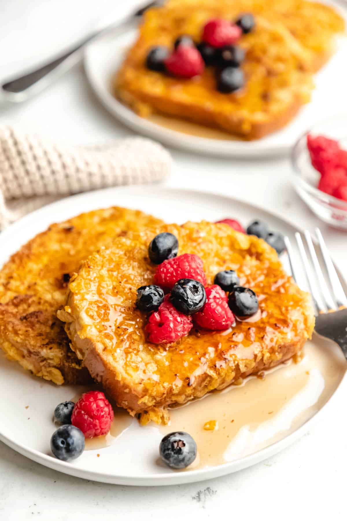 Two pieces of cornflake French toast on a plate in front of another plate of French toast. 