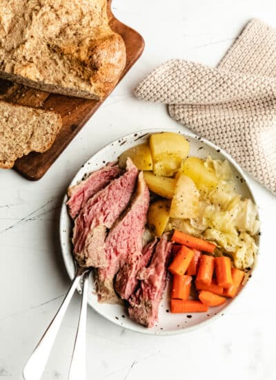 A dish of slow cooker corned beef and cabbage, potatoes, and carrots next to a cutting board with Irish brown bread and a knit napkin.