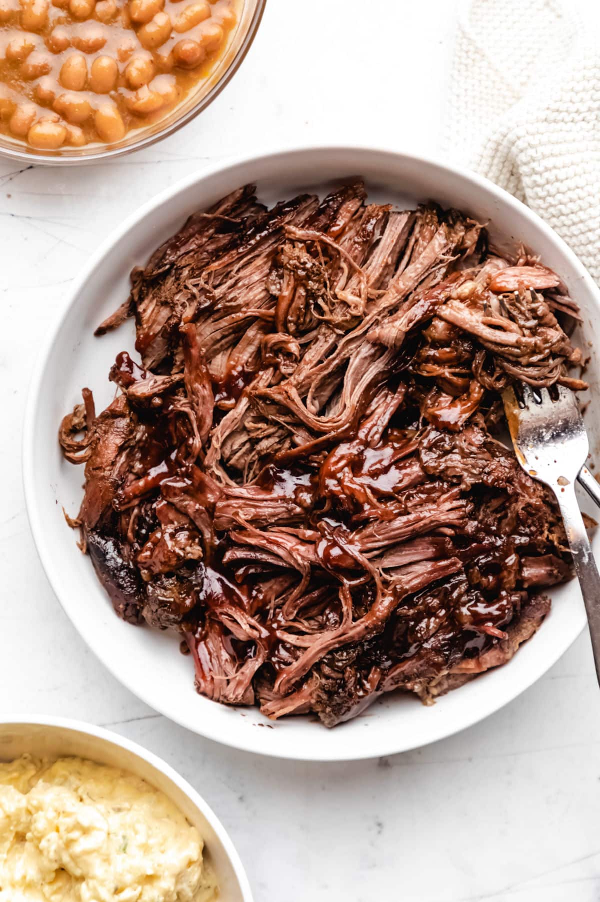 A white plate of shredded slow cooker Texas beef brisket between dishes of baked beans and potato salad. 