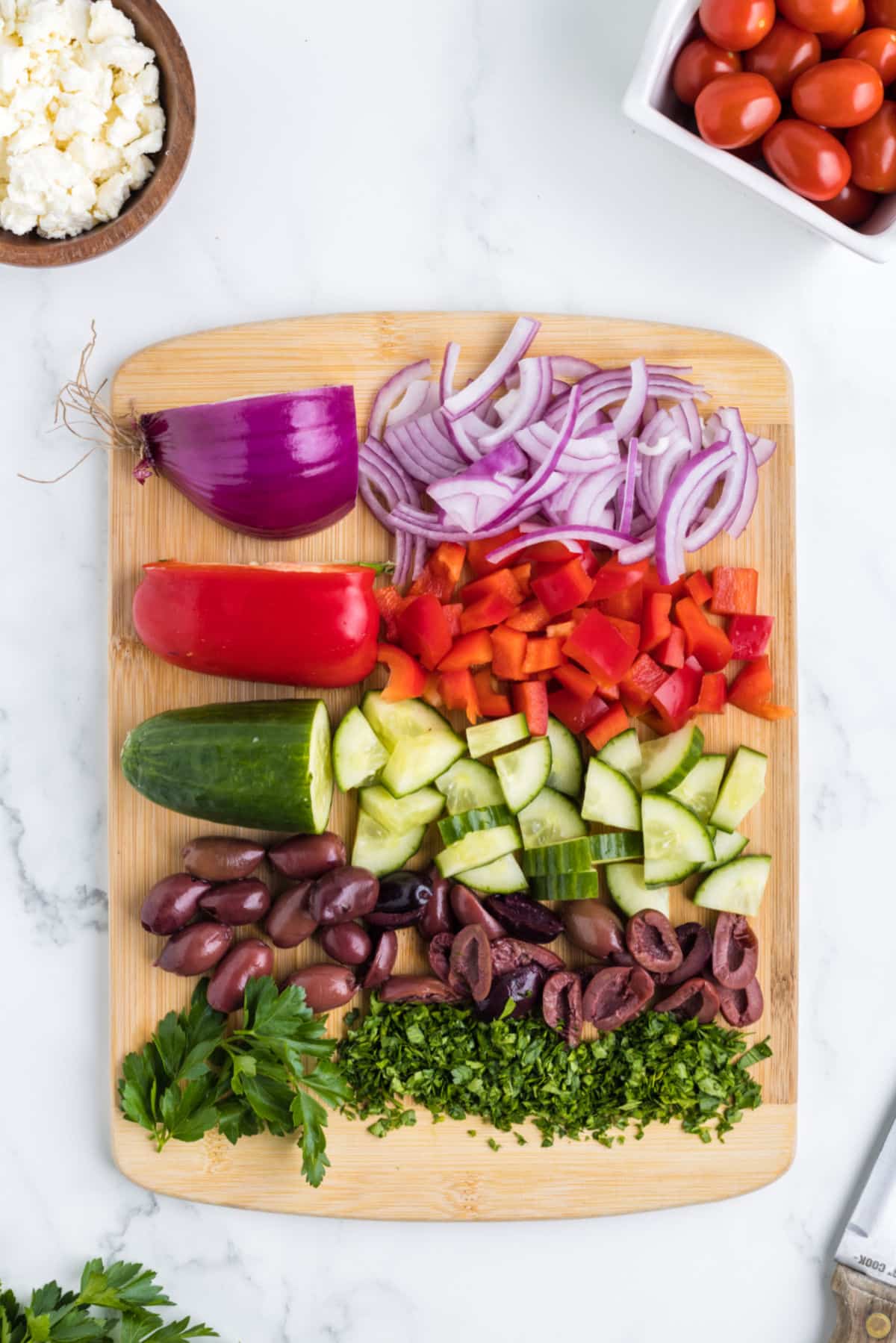 Chopped vegetables for Greek pasta salad on a wooden cutting board.