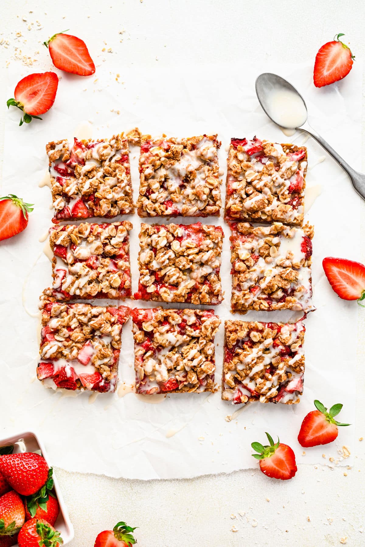 Cut strawberry oatmeal bars on a piece of white parchment paper surrounded by sliced strawberries. 
