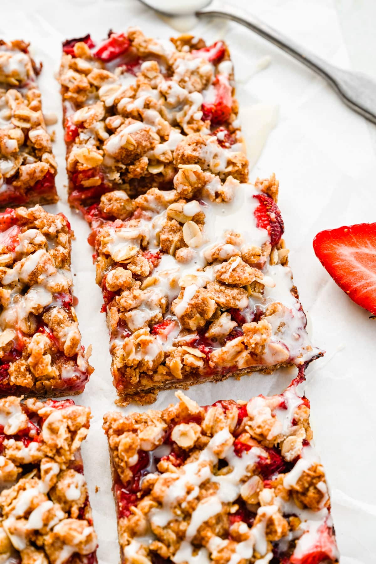 Rows of strawberry oatmeal bars next to a spoon with icing and a sliced strawberry. 