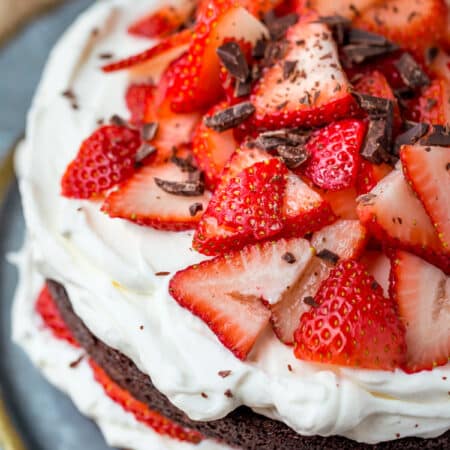 Strawberries and cream chocolate cake on a platter with plates and silverware in the background.