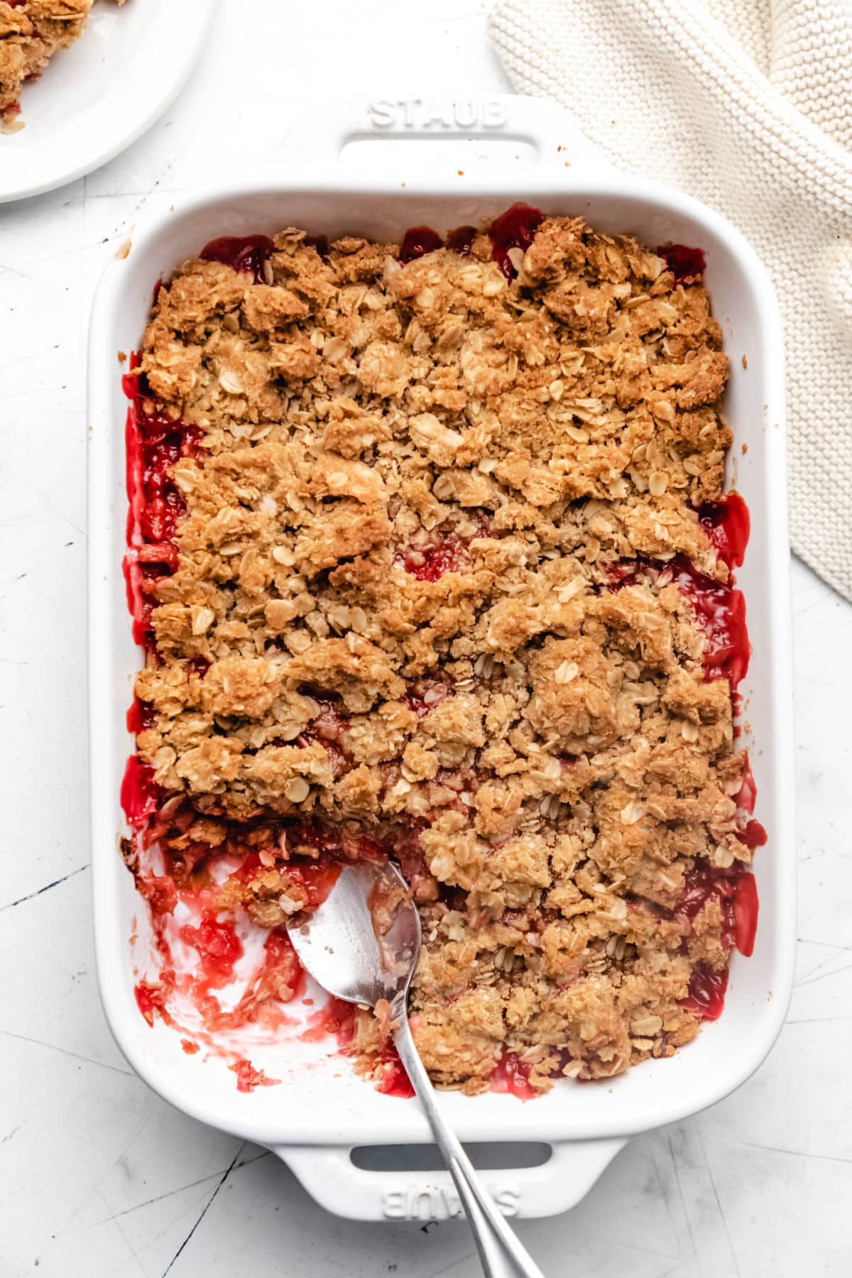 A dish of strawberry rhubarb crisp with part missing and a spoon in it.