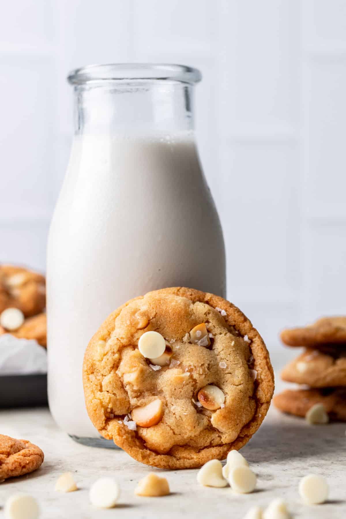 A stack of white chocolate macadamia cookies next to a cookie leaning on a milk bottle.