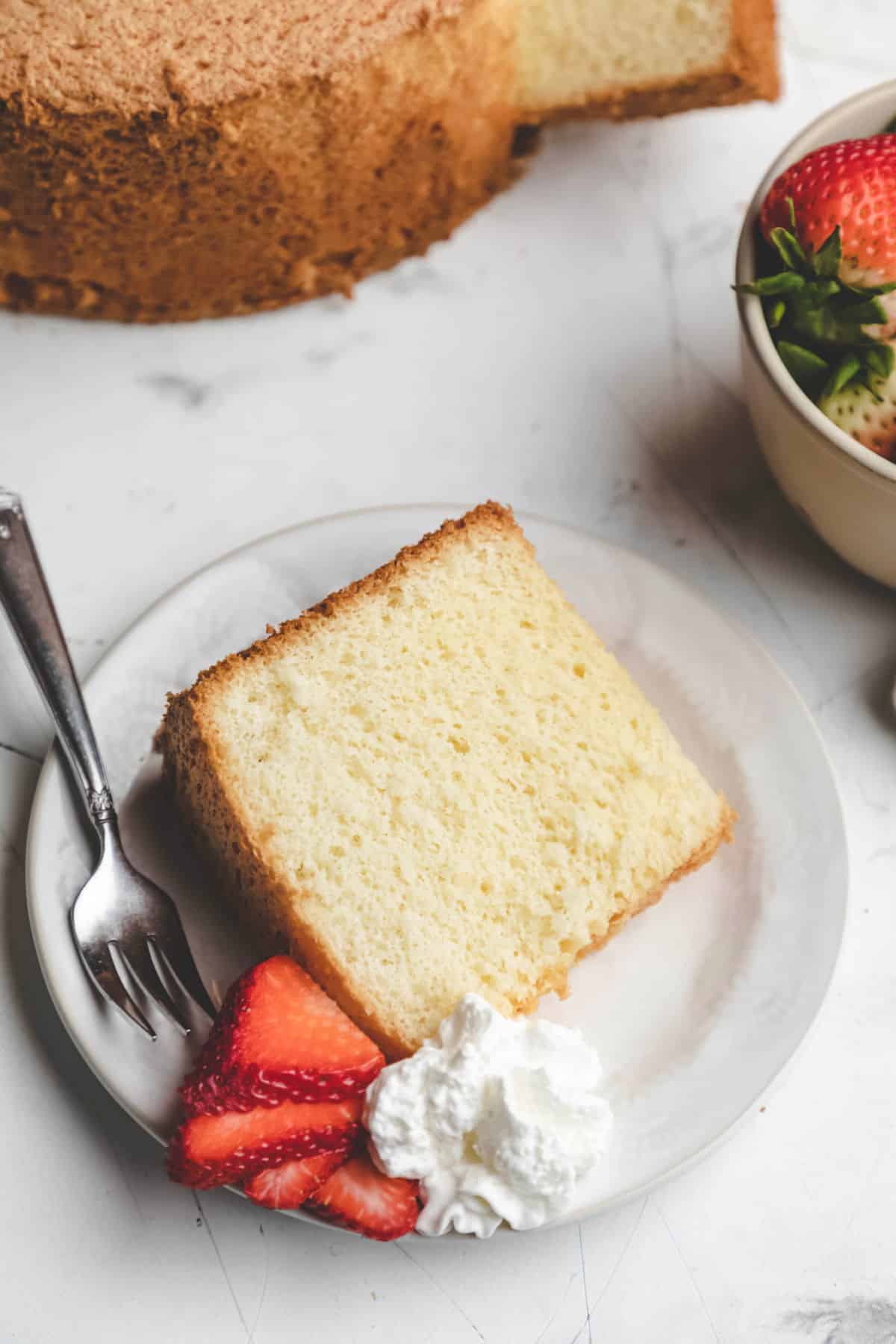 A slice of chiffon cake next to sliced strawberries and whipped cream on a white plate.