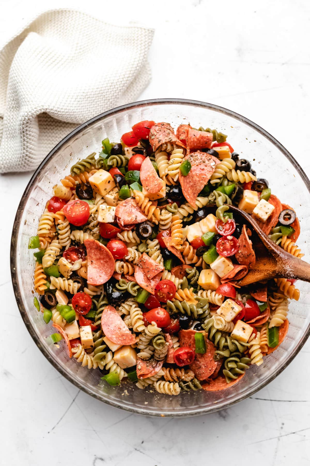 A glass mixing bowl of pasta salad next to a cream knit napkin.