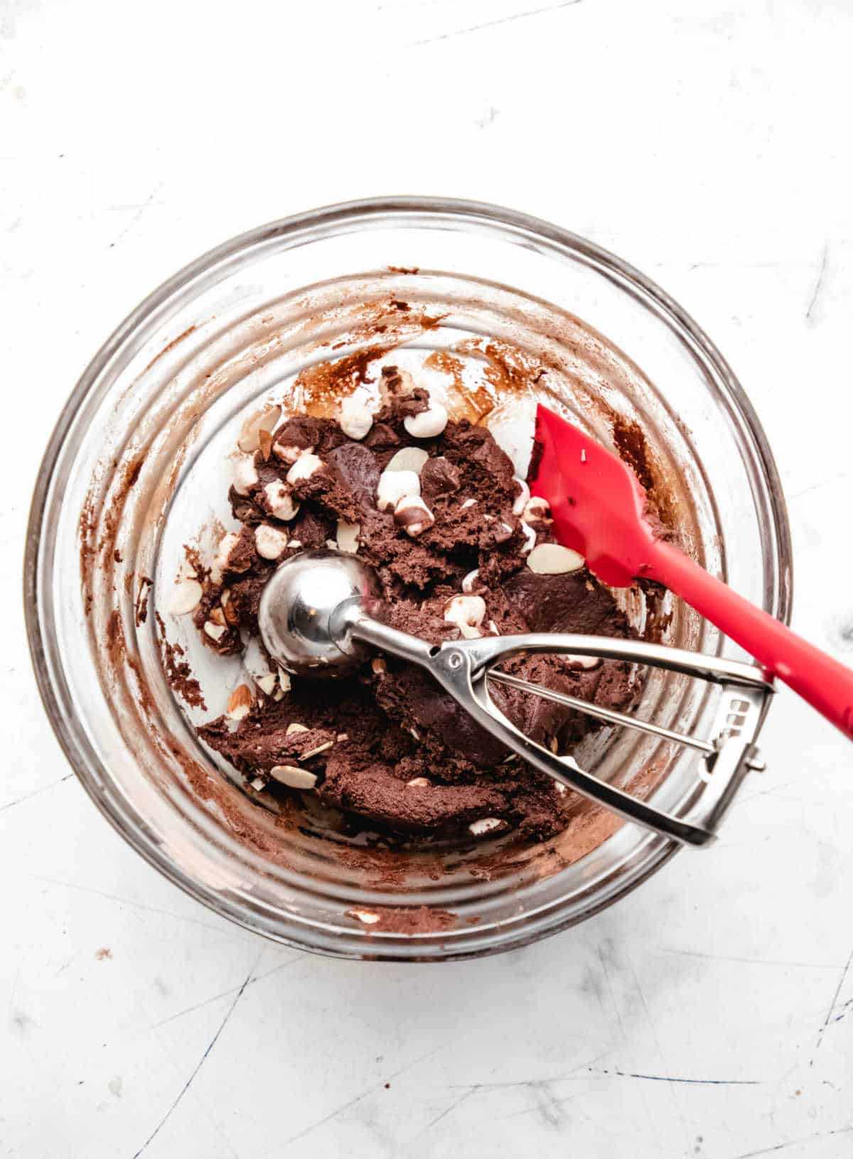 Rocky road cookie dough in a glass mixing bowl with a red spatula and a cookie scoop in it. 