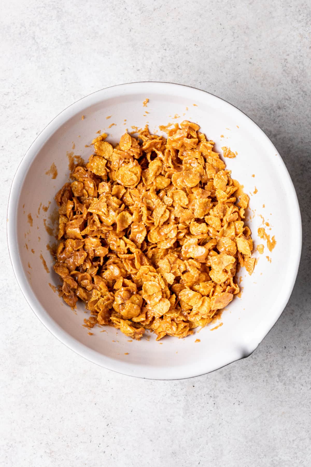 Cornflakes coated in peanut butter mixture in a white mixing bowl. 