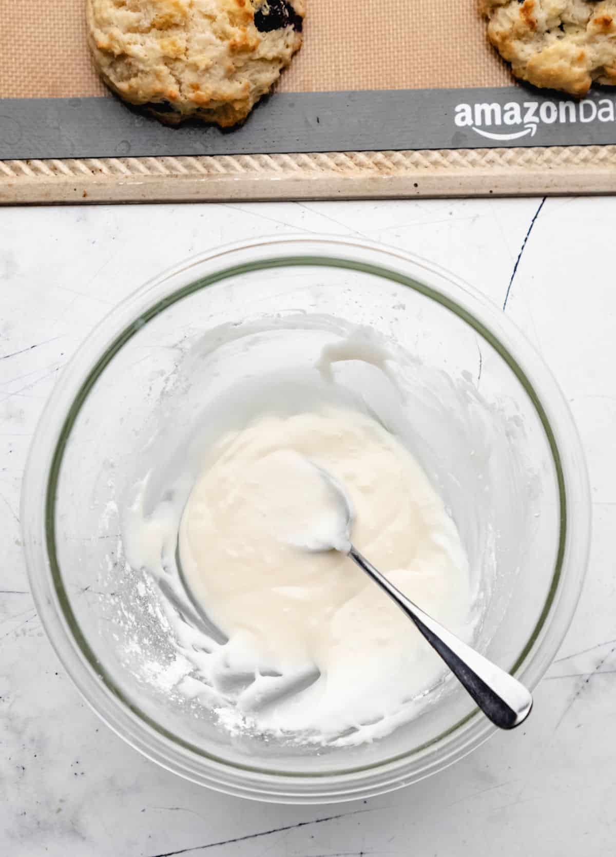 Icing in a glass mixing bowl next to baked blueberry biscuits. 