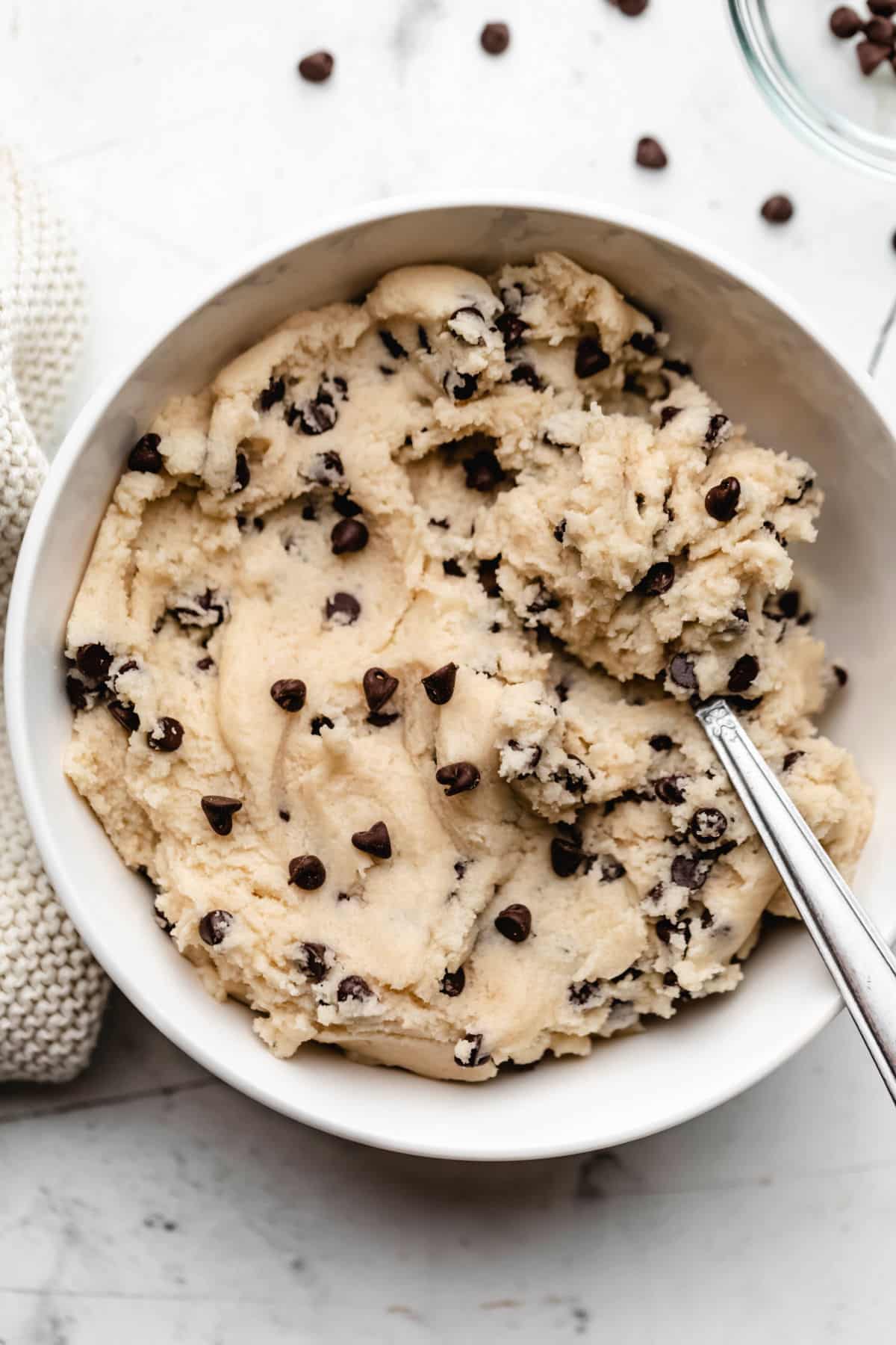A white dish of edible chocolate chip cookie dough with chocolate chips scattered around it.