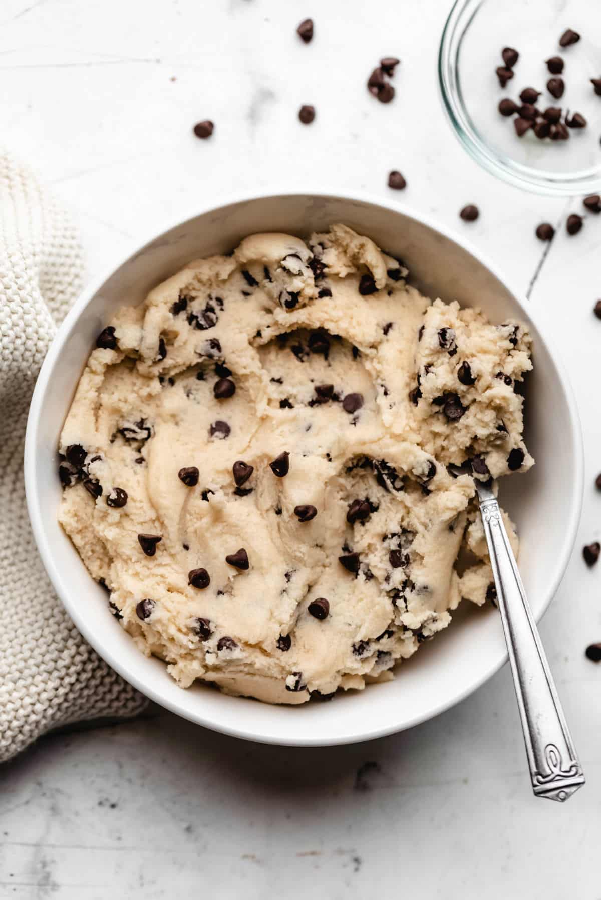 Edible cookie dough in a white bowl next to a dish of chocolate chips. 