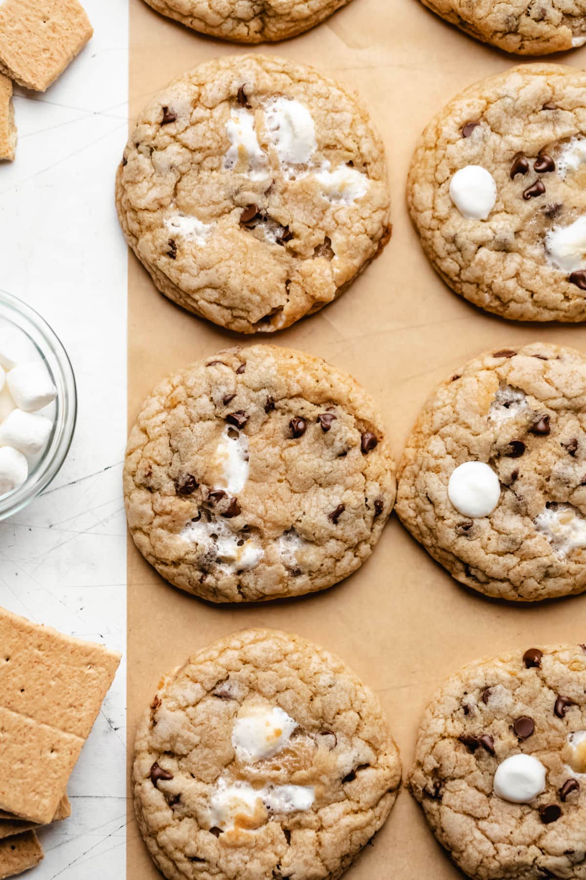 A row of s'mores cookies on brown parchment paper next to a dish of marshmallows.