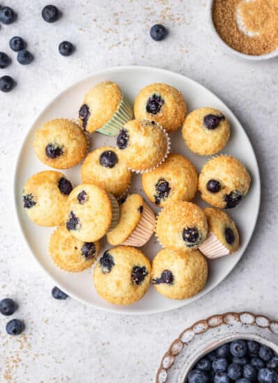 A plate of mini blueberry muffins next to scattered blueberries and a dish of sugar.