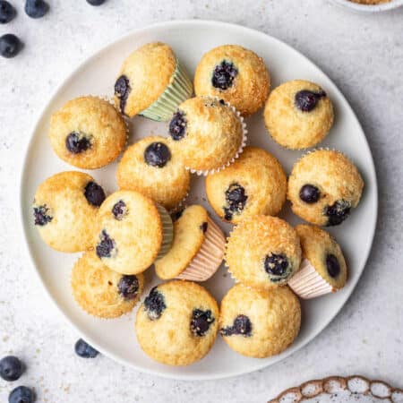 A plate of mini blueberry muffins next to scattered blueberries and a dish of sugar.