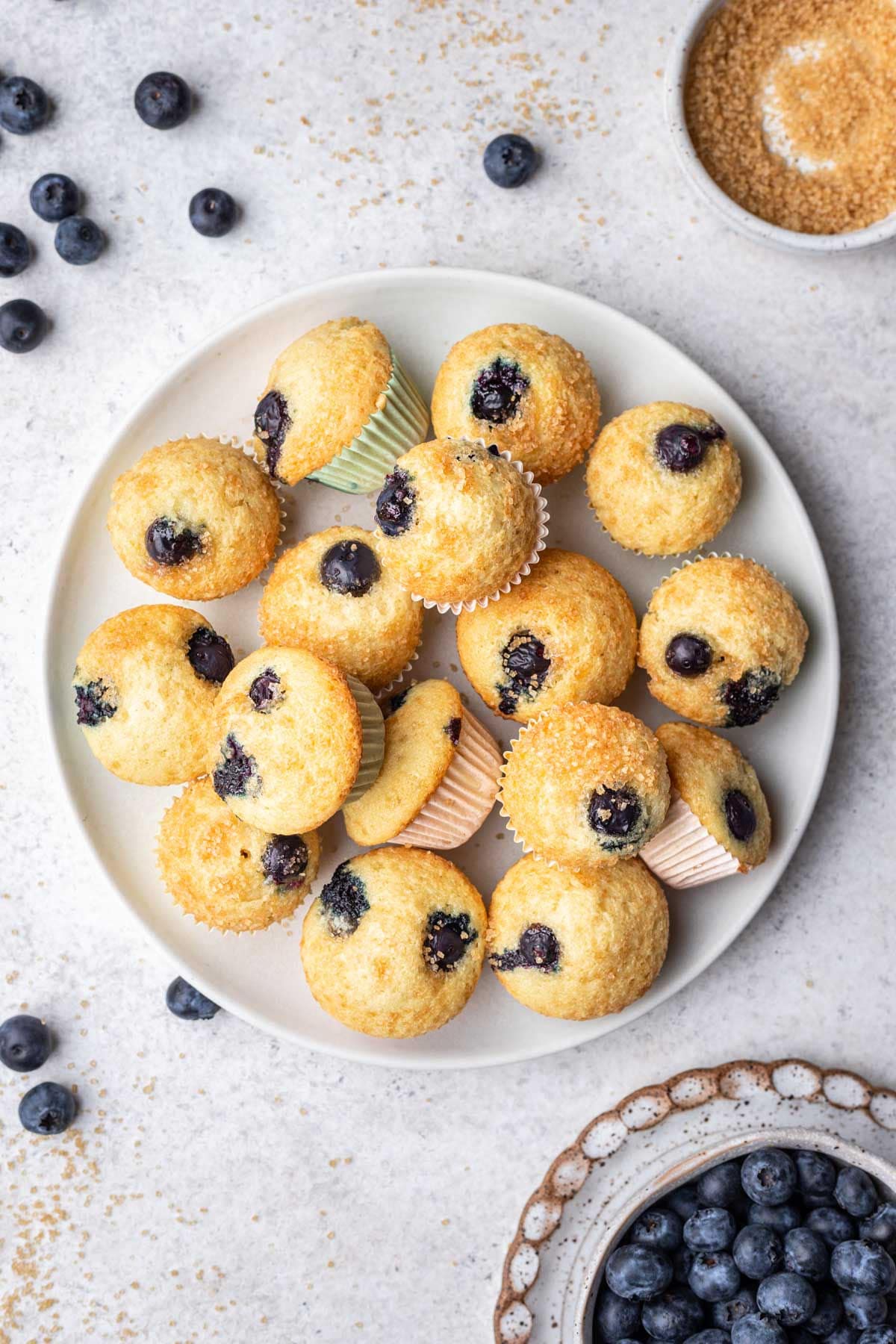 A plate of mini blueberry muffins next to scattered blueberries and a dish of sugar.