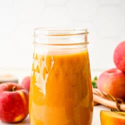 A glass jar of peach syrup surrounded by fresh peaches.