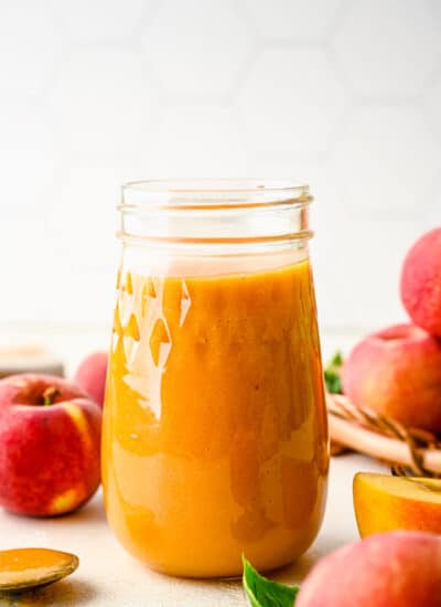 A glass jar of peach syrup surrounded by fresh peaches.