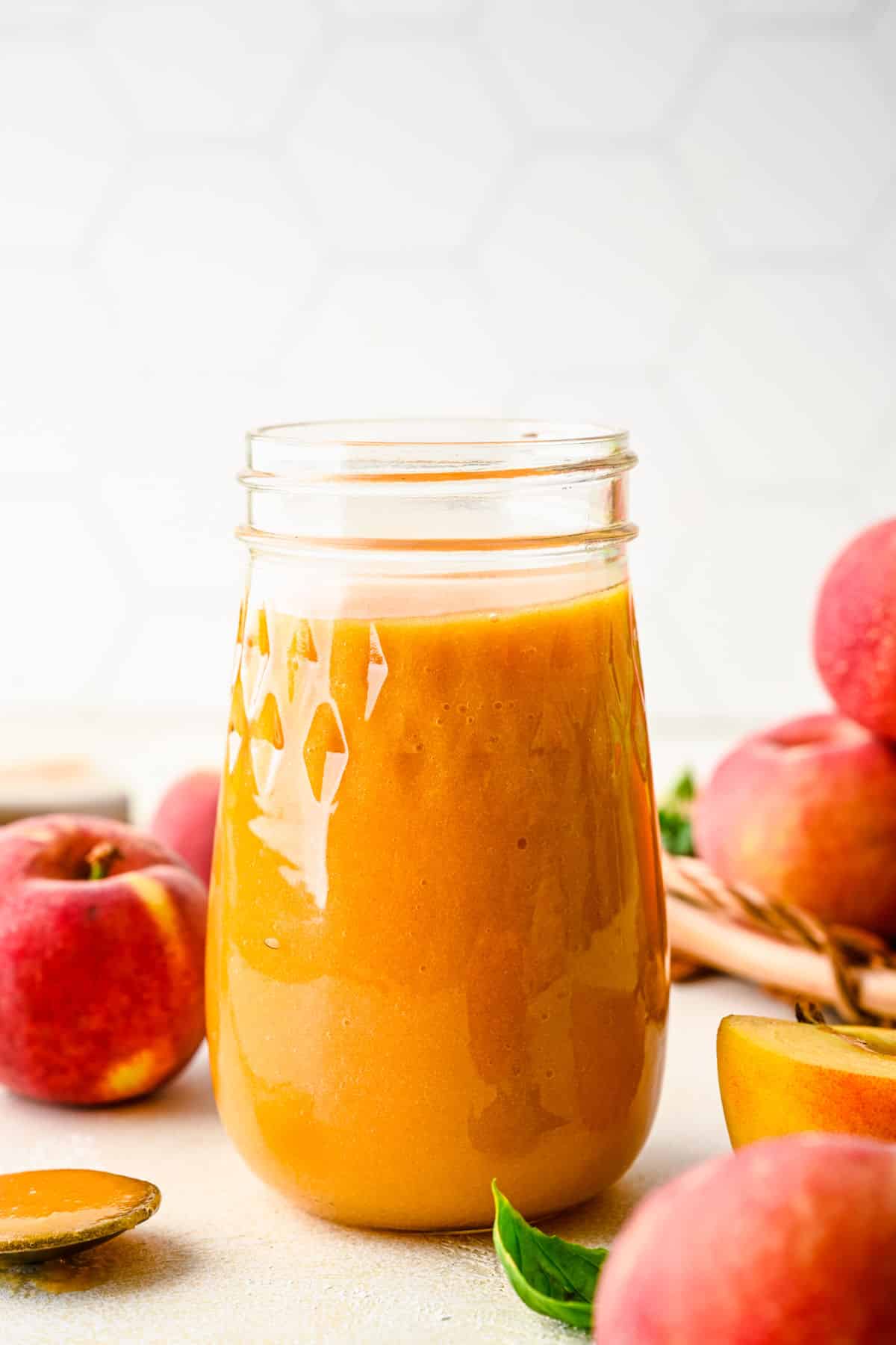 A glass jar of peach syrup surrounded by fresh peaches. 