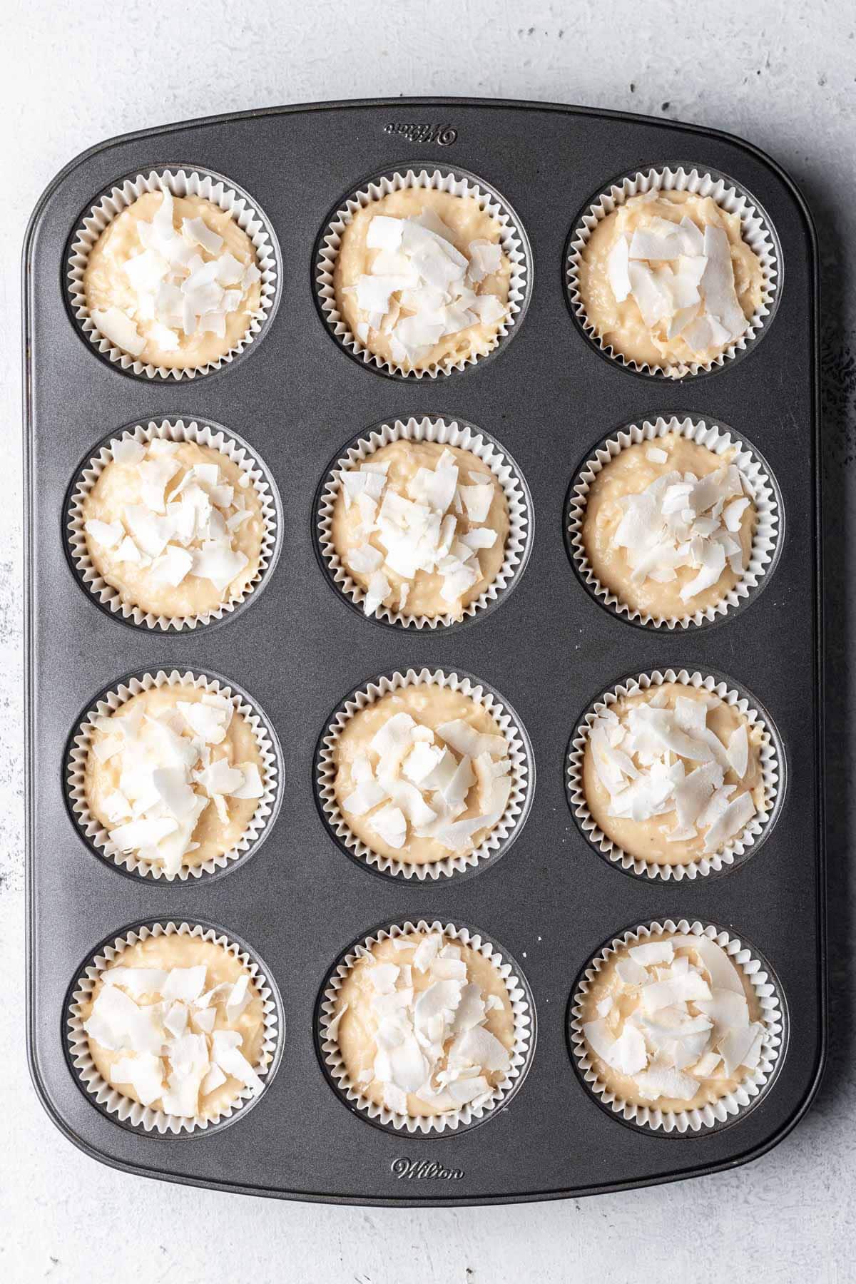 Coconut muffin batter in a muffin tin topped with coconut flakes.