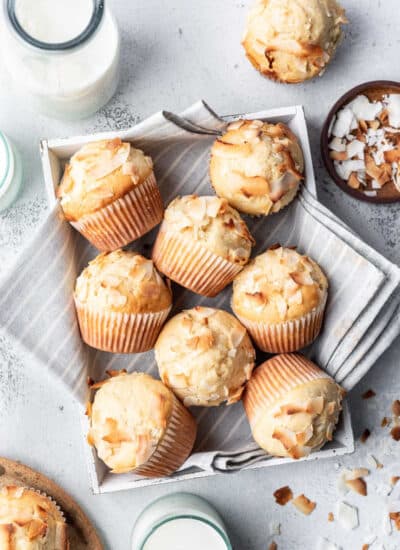 Coconut muffins in a tea towel lined box next to a dish of coconut.