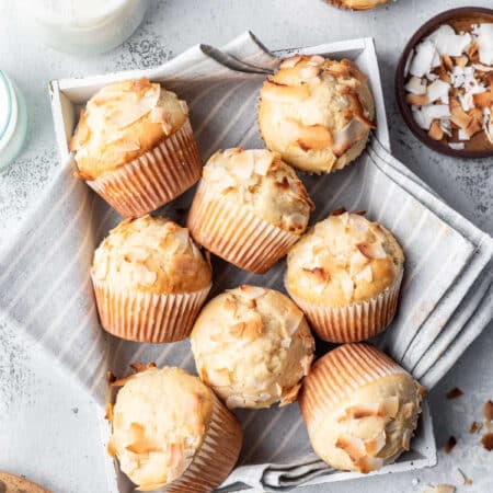 Coconut muffins in a tea towel lined box next to a dish of coconut.