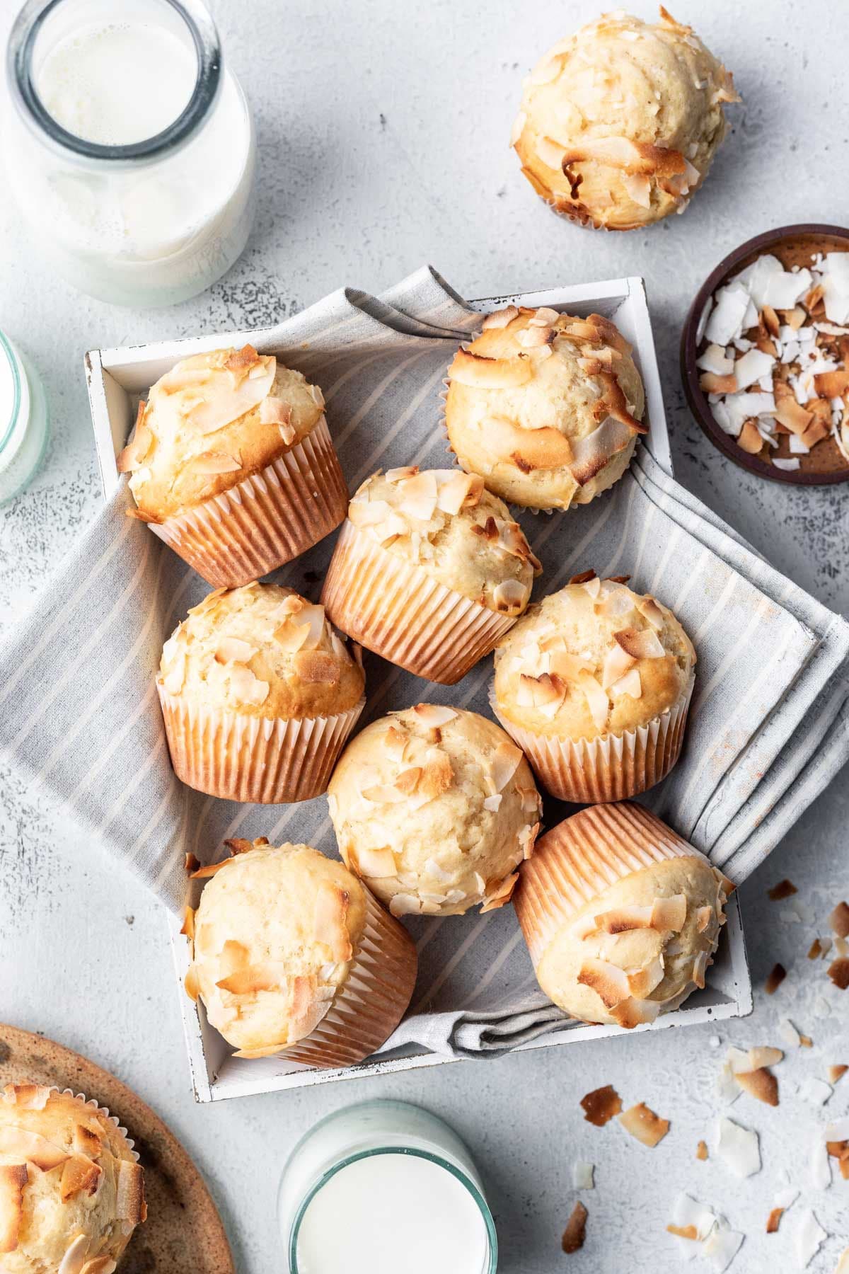 Coconut muffins in a tea towel lined box next to a dish of coconut.