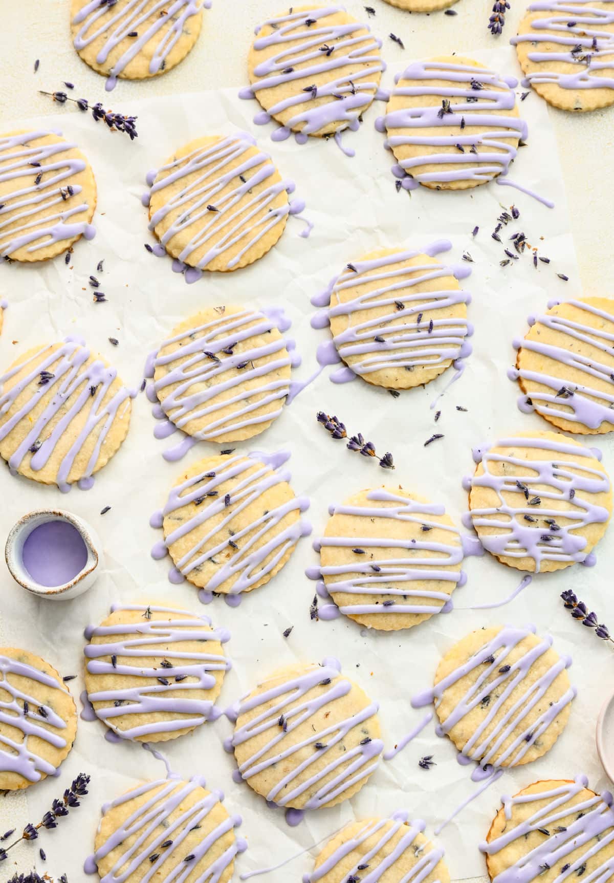 Glazed lavender cookies on a piece of parchment paper surrounded by sprigs of lavender and a dish of glaze.