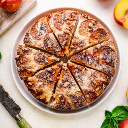 A whole peach cake cut into slices on a white platter surrounded by fresh peaches.