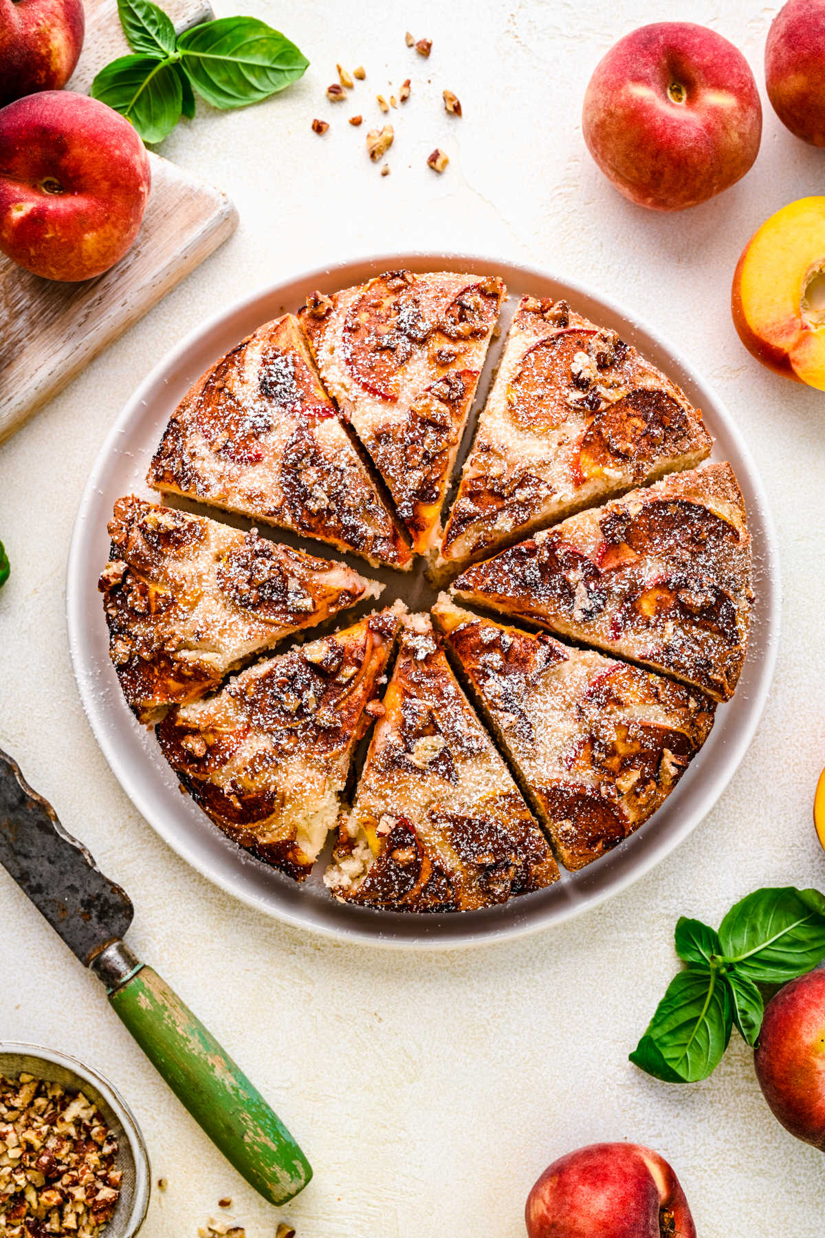 A whole peach cake cut into slices on a white platter surrounded by fresh peaches.