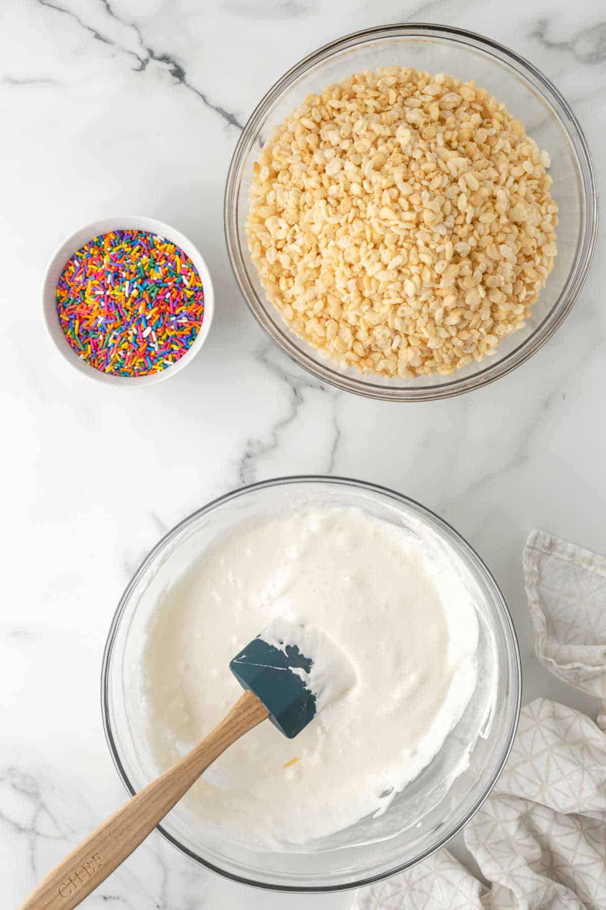 A bowl of melted marshmallow next to bowls of rice krispies cereal and sprinkles. 