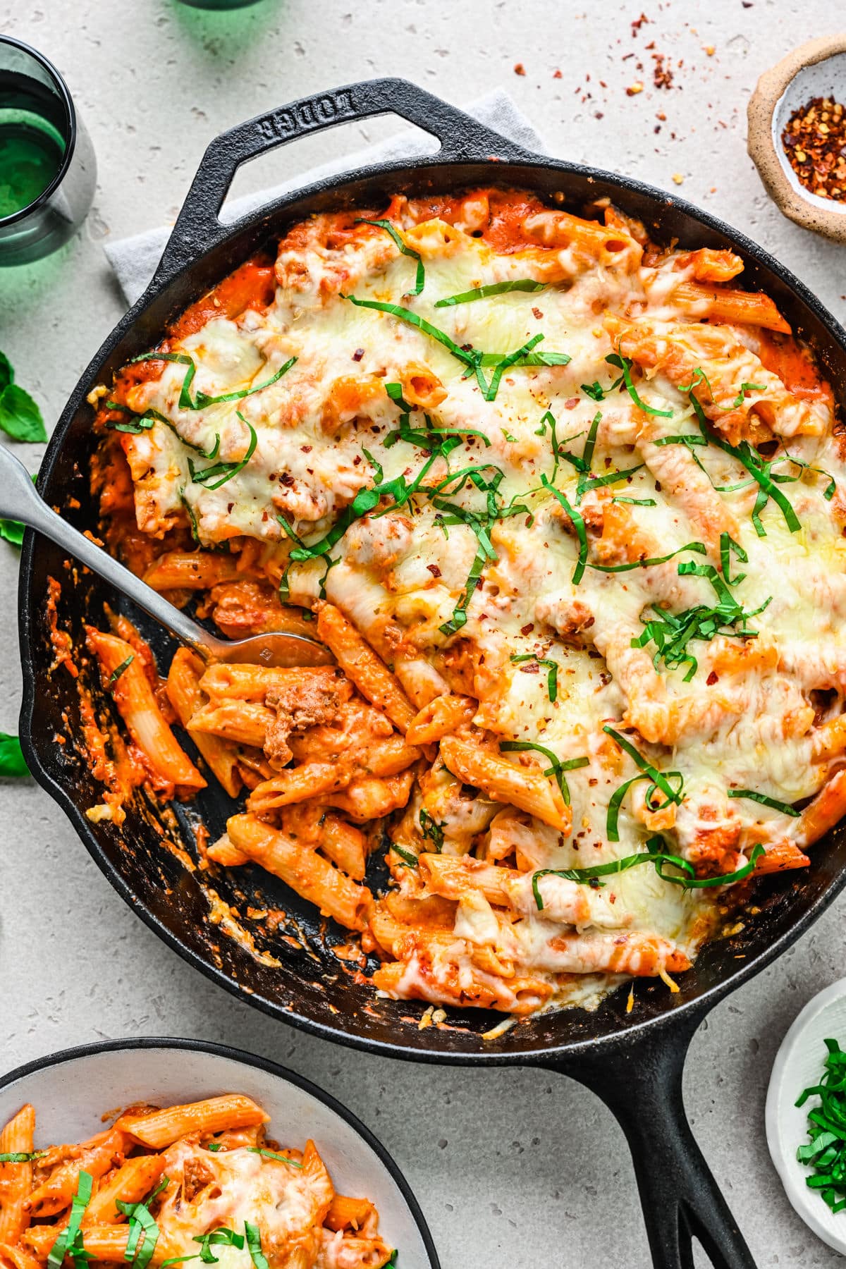 A cast iron skillet filled with baked ziti with a silver spoon scooping up pasta. 