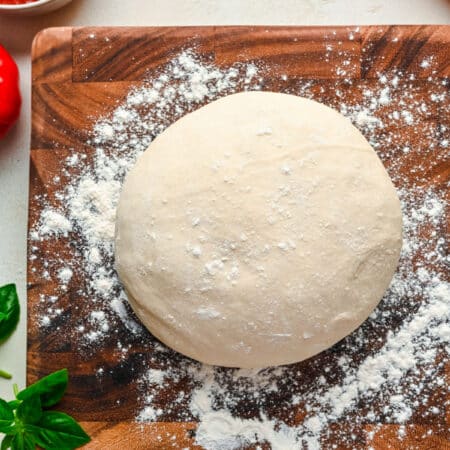 A ball of homemade pizza dough on a wooden cutting board next to a rolling pin.