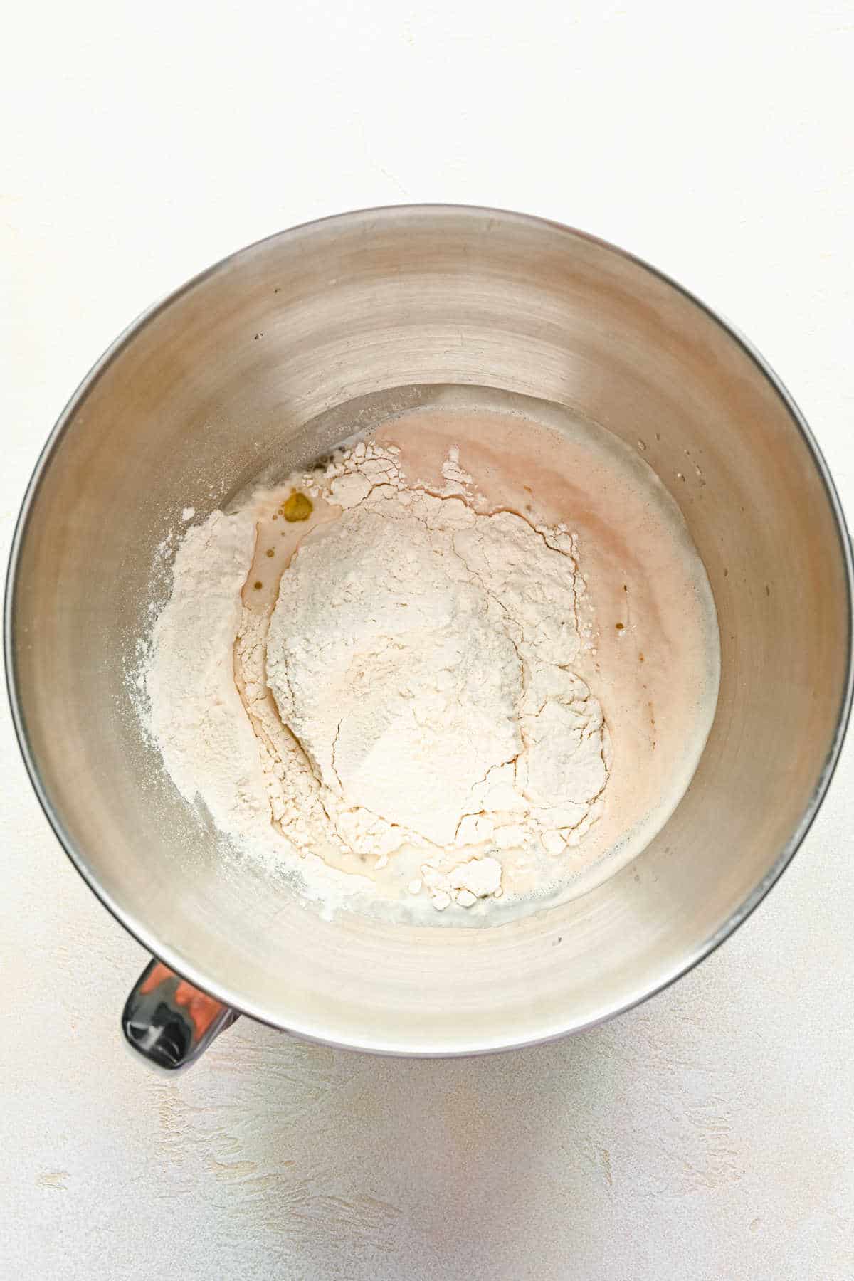 Flour salt and olive oil on top of yeast mixture in a silver mixing bowl.