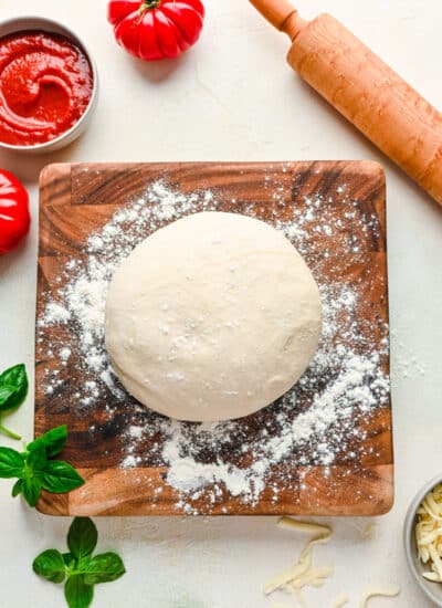 A ball of pizza dough on a wooden cutting board next to a dish of pizza sauce.